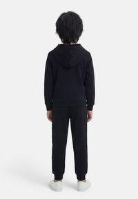 Παιδί που στέκεται με την πλάτη, φορώντας μαύρη μπλούζα με κουκούλα, μαύρο παντελόνι και λευκά αθλητικά παπούτσια, σε απλό φωτεινό φόντο.