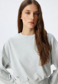 Hellgrauer gerippter Pullover mit Rundhalsausschnitt, überschnittenen Schultern und elastischem Saum, der eine weiche Textur aufweist. Die Haare fallen in sanften Wellen.