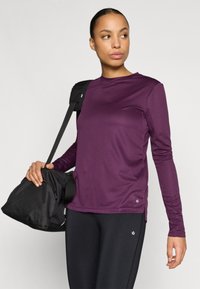 Pitkähihainen, violetti urheilupaita, joka on valmistettu sileästä kankaasta, yhdistettynä mustaan laukkuun. Paidassa on pyöreä kaula-aukko ja hienovaraiset sivuhalkiot.