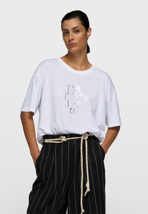 Biała koszulka z krótkim rękawem z błyszczącym srebrnym wzorem na przodzie. W połączeniu z czarno-białymi spodniami w paski, z beżowym paskiem.