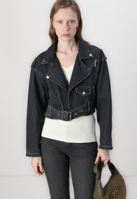 Kuttet svart denimjakke med sølvfargede detaljer, frontglidelås og flere lommer; båret over en lyse farget topp, kombinert med mørke jeans.