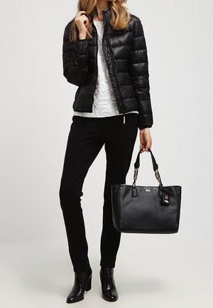 Femme portant une doudoune noire, un haut blanc à motifs, un pantalon noir et des bottines, tenant un grand sac à main en cuir noir par ses poignées.