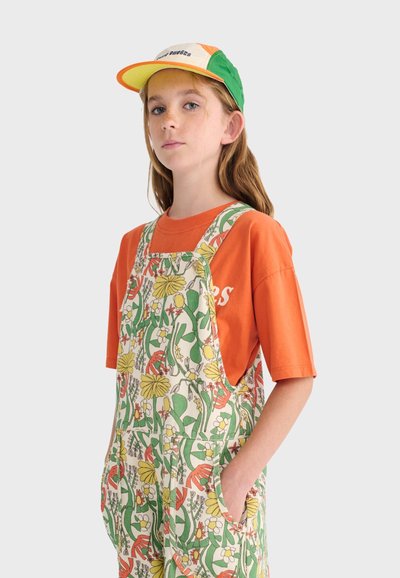 Jeune personne portant une casquette verte, orange et jaune, un t-shirt orange et une salopette à motif floral, les mains dans les poches, sur un fond uni.