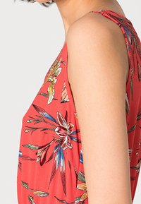 Robe sans manches fleurie en tissu rouge corail, présentant des motifs de fleurs tropicales vibrantes et une coupe décontractée, avec une couture d'épaule visible et une texture.