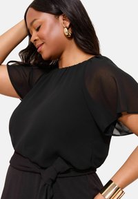 Schwarze Bluse mit durchsichtigen kurzen Ärmeln, rundem Ausschnitt und einer gebundenen Taillendetail. Ein goldenes Armreif sorgt für Kontrast und betont den Look.