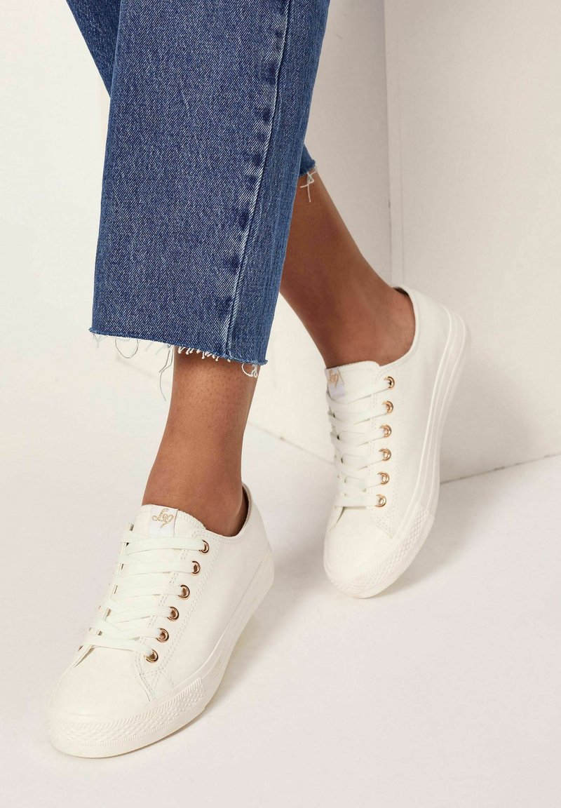 Sneakers bianchi con occhielli dorati, punta testurizzata e tomaia liscia, abbinati a jeans blu cropped e strappati, che mostrano una vestibilità rilassata.