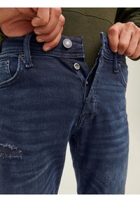 Jeans i mörkblå denim med en lätt sliten textur och synlig sömnad. Har en metallknapp och dragkedja, med sidfickor.