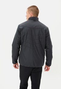 Homme portant une veste matelassée noire et un pantalon noir, se tenant de dos face à l'appareil photo sur un fond blanc.