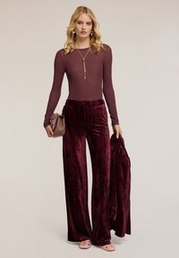 Bordeaux-velours weite Hose, kombiniert mit einem taillierten Langarmoberteil, accessoriert mit einer goldfarbenen Halskette und einer kleinen braunen Handtasche.