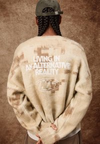 Krämfärgad oversized tröja med en mjuk, strukturerad yta, med bruna abstrakta mönster och vit broderad text på baksidan.