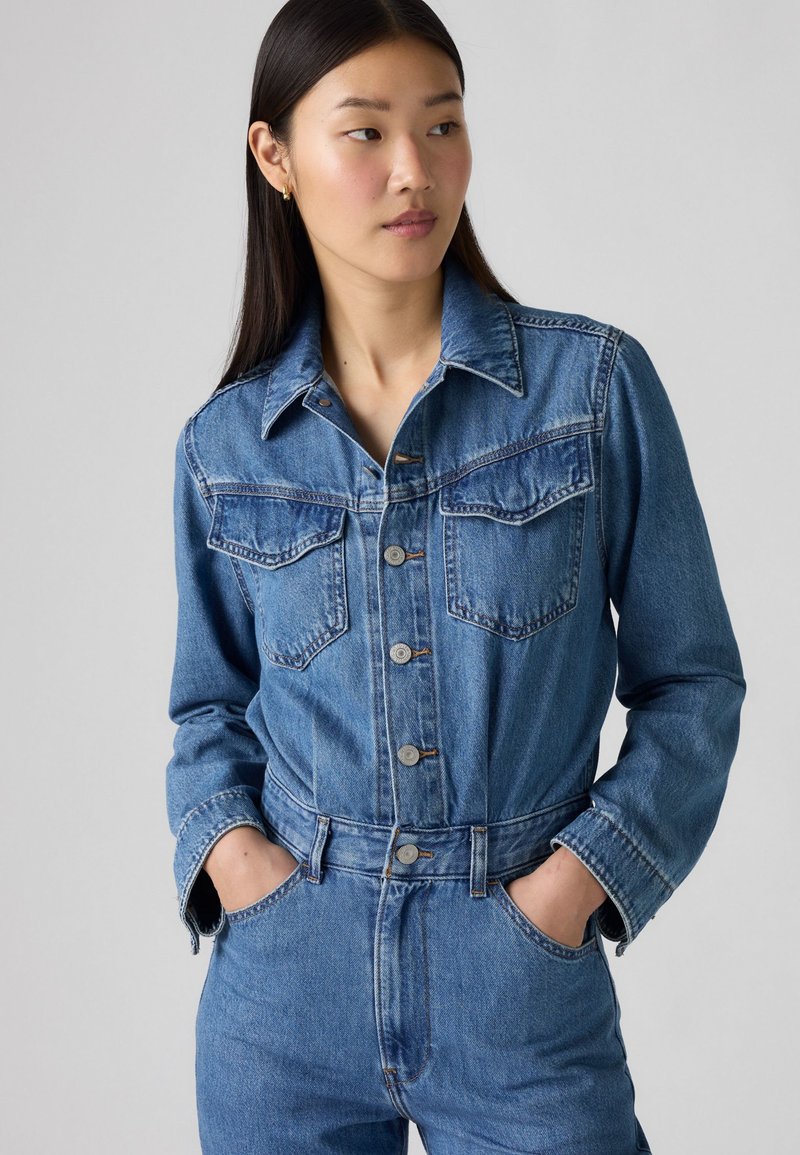 Combinaison en denim bleu moyen. Comprend une fermeture à boutons sur le devant, deux poches poitrine et des manches longues avec poignets boutonnés. Coupe ajustée et jambes fuselées.