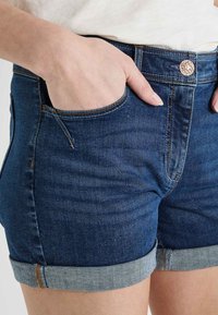 Pantalones cortos de mezclilla en azul oscuro con dobladillo, que presentan un bolsillo frontal, costuras de cobre y un cierre de botón metálico.