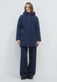 Donna che indossa un cappotto imbottito blu navy con cappuccio e zip frontale, pantaloni larghi blu navy e scarpe nere, in piedi su uno sfondo bianco.