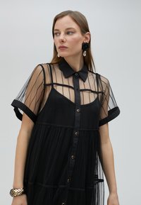 Robe noire avec un corsage en tulle rayé et un col. Jupe en tulle en dessous et boutons dorés. Manches courtes roulées.