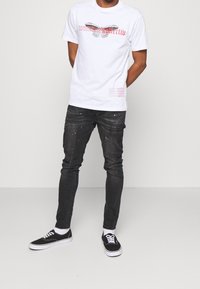 Camiseta blanca de algodón con un gráfico de mariposa y texto en el frente; combinada con unos vaqueros oscuros con salpicaduras de pintura y zapatillas negras.