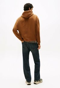 Sweatshirt à capuche marron avec une finition texturée, associé à un jean bleu foncé et des baskets à motifs. Le modèle fait face à l'opposé de l'appareil photo.