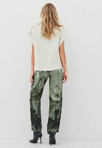 Woman with wavy blonde hair wearing a white knitted sleeveless top, green abstract-print pants, and black heeled ankle boots, facing away.