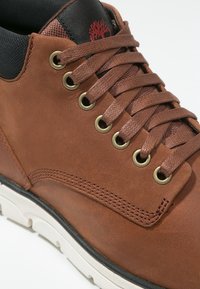 Chaussure en cuir marron avec œillets dorés, lacets marron et un col noir texturé. La semelle est blanche avec une bande d'accent noire.