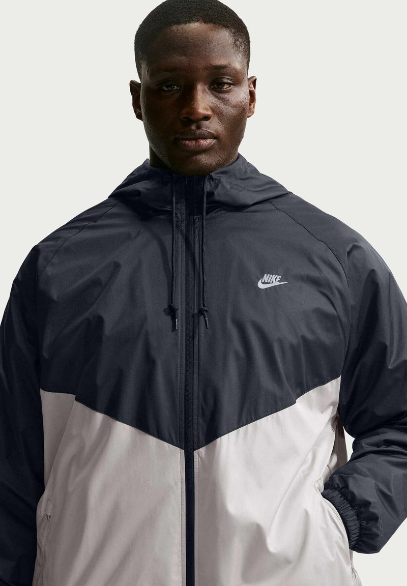 Man draagt een zwart-witte Nike windbreaker met capuchon en handen in de zakken, kijkend naar voren tegen een effen achtergrond.