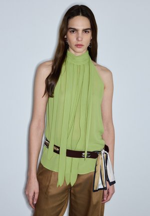 Vrouw met een mouwloze limoengroene blouse met sjaalkraagdetail en een bruine broek, staand tegen een effen lichtgrijze achtergrond.