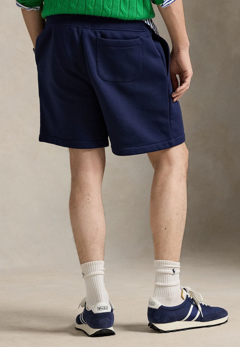 Navy blue athletic shorts with a smooth texture, featuring a back pocket and contrasting stitch accents. Paired with navy sneakers and white socks.