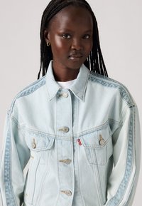 Chaqueta de mezclilla azul claro con un corte relajado, grandes bolsillos, cierre de botones y detalles de costura en contraste. Presenta una distintiva marca de Levi's.