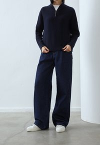 Marineblauer gerippter Strickpullover mit Viertelreißverschluss, kombiniert mit locker sitzenden dunklen Jeans und weißen Sneakern, stehend auf einem Betonboden.