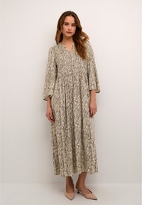 Long, flowing dress in light beige featuring a wavy black pattern. Smocked bodice and three-quarter sleeves. Paired with nude heeled shoes.