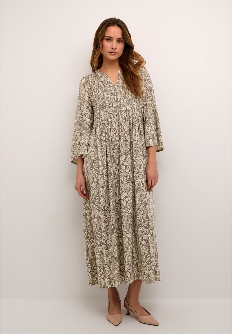 Long, flowing dress in light beige featuring a wavy black pattern. Smocked bodice and three-quarter sleeves. Paired with nude heeled shoes.