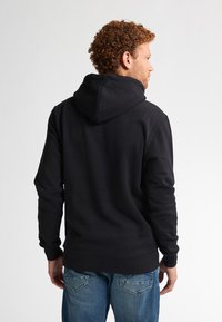 Felpa nera con cappuccio regolabile, tessuto a trama, tasca a marsupio e polsini a coste. Indossata con jeans blu, vista dal retro.