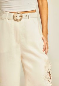 Wijde crème broek met kantdetails aan de zijkant. Gecombineerd met een textuurstof en een geweven, ronde gesp in een lichte tint.