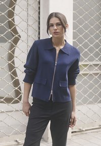 Marineblaue Zipjacke mit Kragen, Dreiviertelärmeln und zwei vorderen Taschen. Der Stoff wirkt strukturiert und warm.