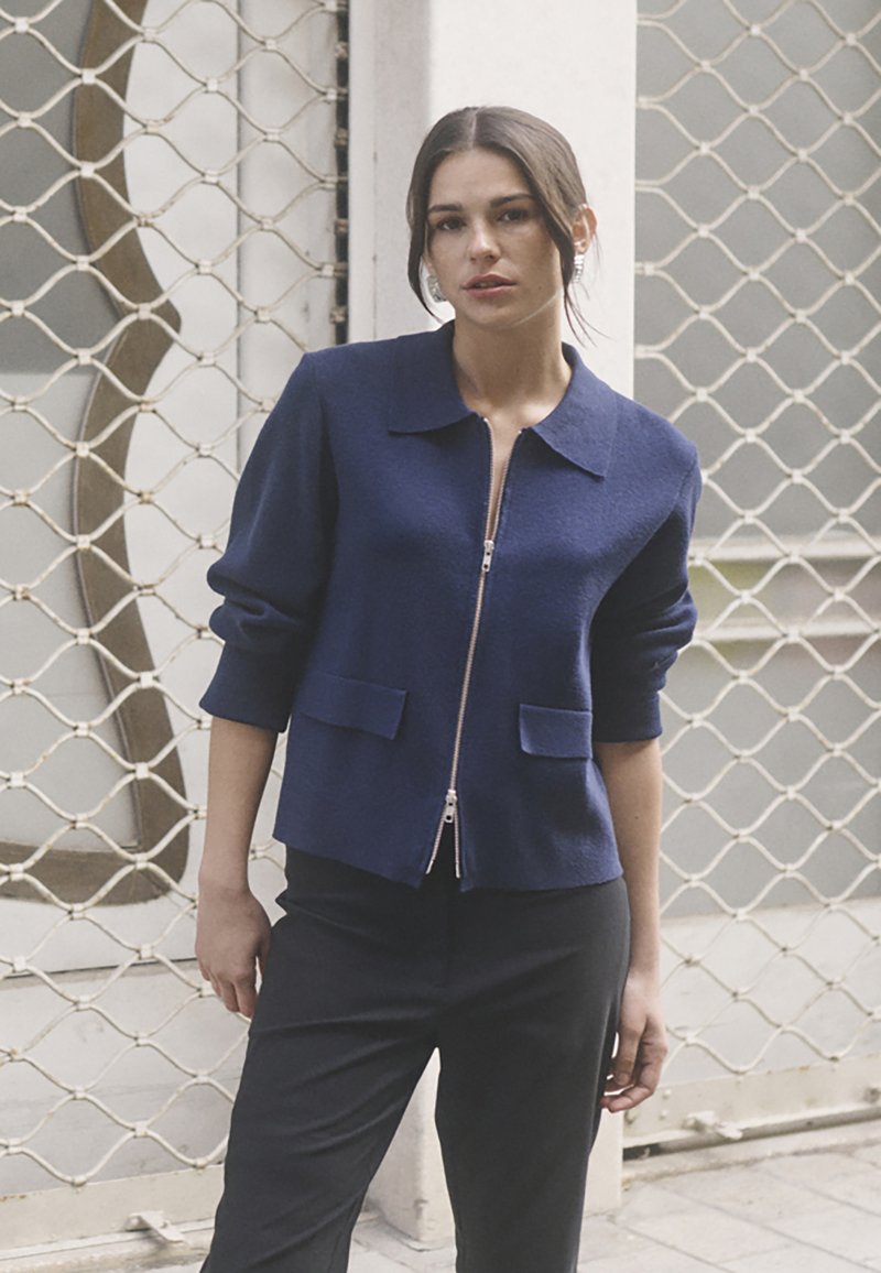 Marineblaue Zipjacke mit Kragen, Dreiviertelärmeln und zwei vorderen Taschen. Der Stoff wirkt strukturiert und warm.