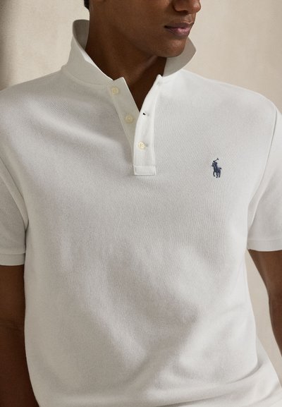 Mężczyzna w białej koszulce polo z podniesionym kołnierzykiem i trzema guzikami, z małym ciemnym haftowanym logo konia na piersi.