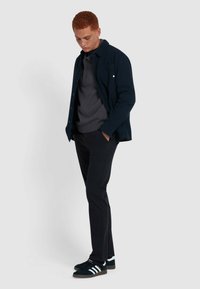 Marineblauwe knoop-jack over een grijze polo, zwarte broek en donkere sneakers met witte strepen, met een textuurstof en een casual pasvorm.