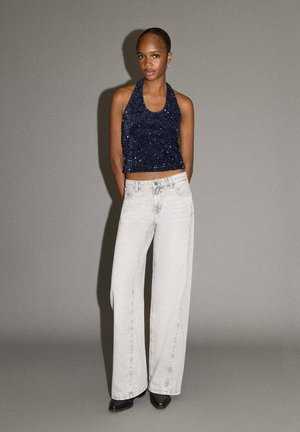 Top halter blu con paillettes abbinato a jeans a gamba larga grigio chiaro e stivaletti neri con punta. Texture lisce e vestibilità ampia in entrambi i capi.
