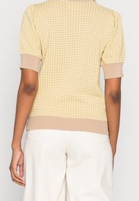 DZIANINOWY TOP w jasnym żółtym kolorze z fakturowanym wzorem, krótkimi rękawami oraz beżowym ściągaczem u dołu i przy mankietach. Noszony z jasnymi spodniami.