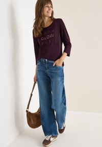 Camicia a maniche lunghe viola scuro con testo in rilievo, abbinata a jeans blu larghi e sneakers marroni. Porta una borsa a spalla marrone.