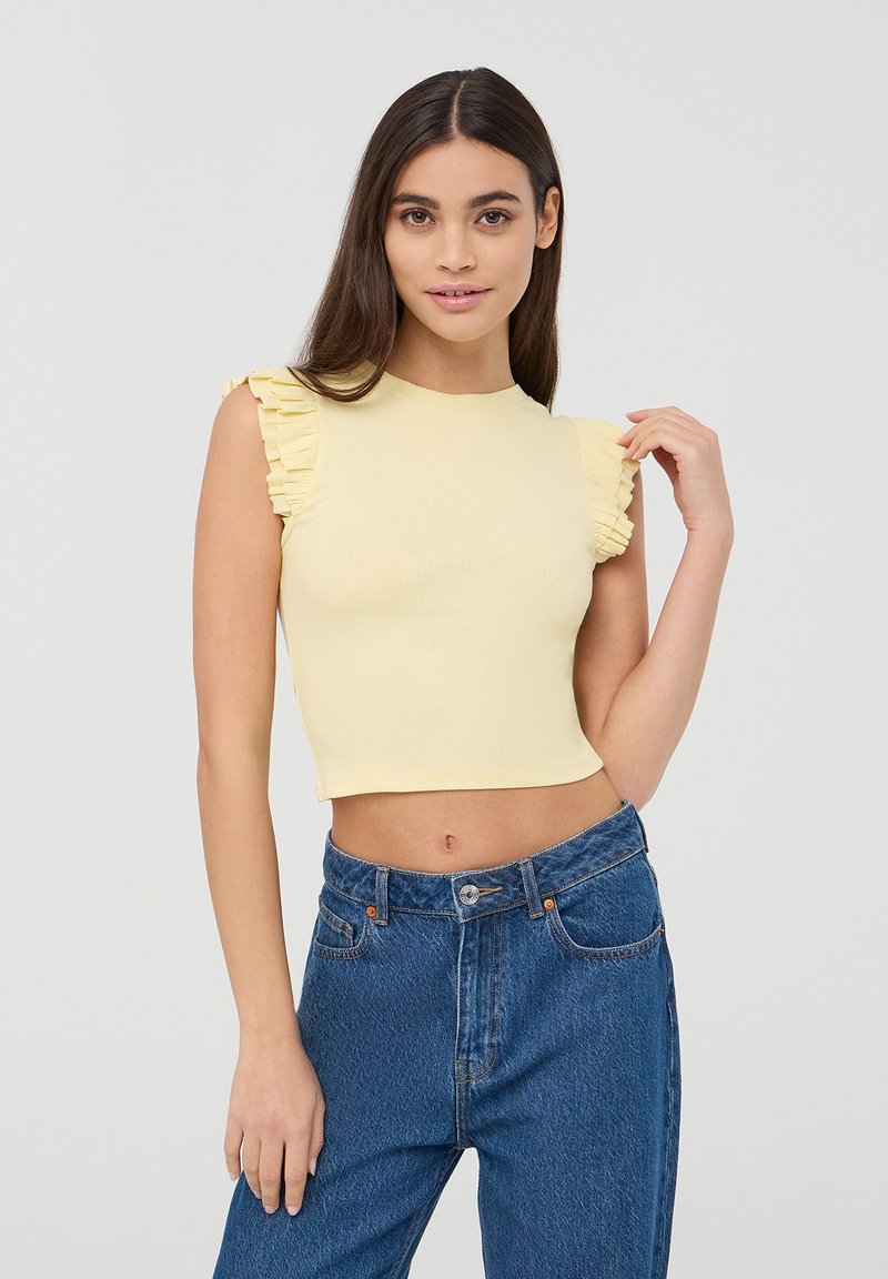 Cropped top di un leggero giallo con texture a coste, con dettagli di maniche a volant, abbinato a jeans in denim blu a vita alta. Design semplice, senza motivi.