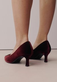 Bordo velveta augstpapēžu kurpes ar norādītu purngalu un nelielu sudraba dekoratīvu akcentu. Tām ir šaurējs papēdis un maiga tekstūra.