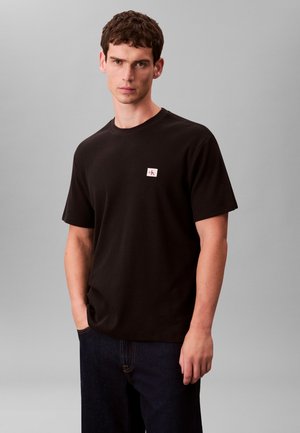 Mladý muž s krátkymi hnedými vlasmi v jednoduchom čiernom tričku s krátkymi rukávmi a malým bielo-červeným logom na hrudi, stojaci pred sivým pozadím.