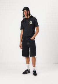Black cotton t-shirt with graphic design on the chest, paired with knee-length black denim shorts and black sneakers with white stripes.