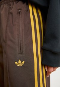 Pantaloni sportivi maro cu dungi galbene, buzunar lateral cu fermoar și un logo brodat. Textură moale cu un design conic.
