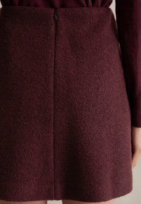 Bordowa sukienka z wełny o teksturowanym wykończeniu, z centralnym szwem i dyskretnym zamkiem na plecach. Długie rękawy dopełniają projekt.