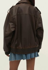 Brun imitation læderjakke med krave, ribbet nederste kant og skulderepauletter, parret med en lys denimnederdel.