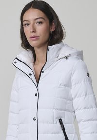 Chaqueta puffer blanca con diseño de acolchado vertical, detalles de cremallera negra y capucha desmontable con un suave forro interior.