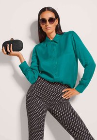 Vrouw in een turquoise knopenblouse en zwarte gemêleerde broek, met een zwarte clutch in de hand, ronde zonnebril op en ringoorbellen.