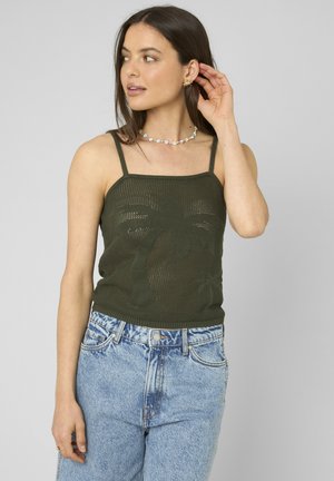 Jeune femme aux longs cheveux foncés portant un débardeur en maille vert foncé avec un motif de palmier et un jean taille haute bleu clair, ajustant sa boucle d’oreille.