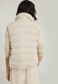 Jasnobeżowa pikowana kamizelka z wysokim kołnierzem, poziomym przeszyciem i regulowanymi bocznymi sznureczkami. Materiał wydaje się miękki i gładki.