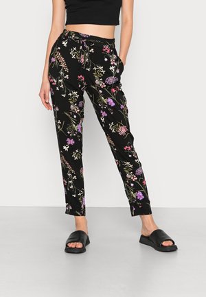 Vrouw die zwarte broek met bloemenprint draagt, met een trekkoord in de taille en zwarte slippers, staand tegen een witte achtergrond.